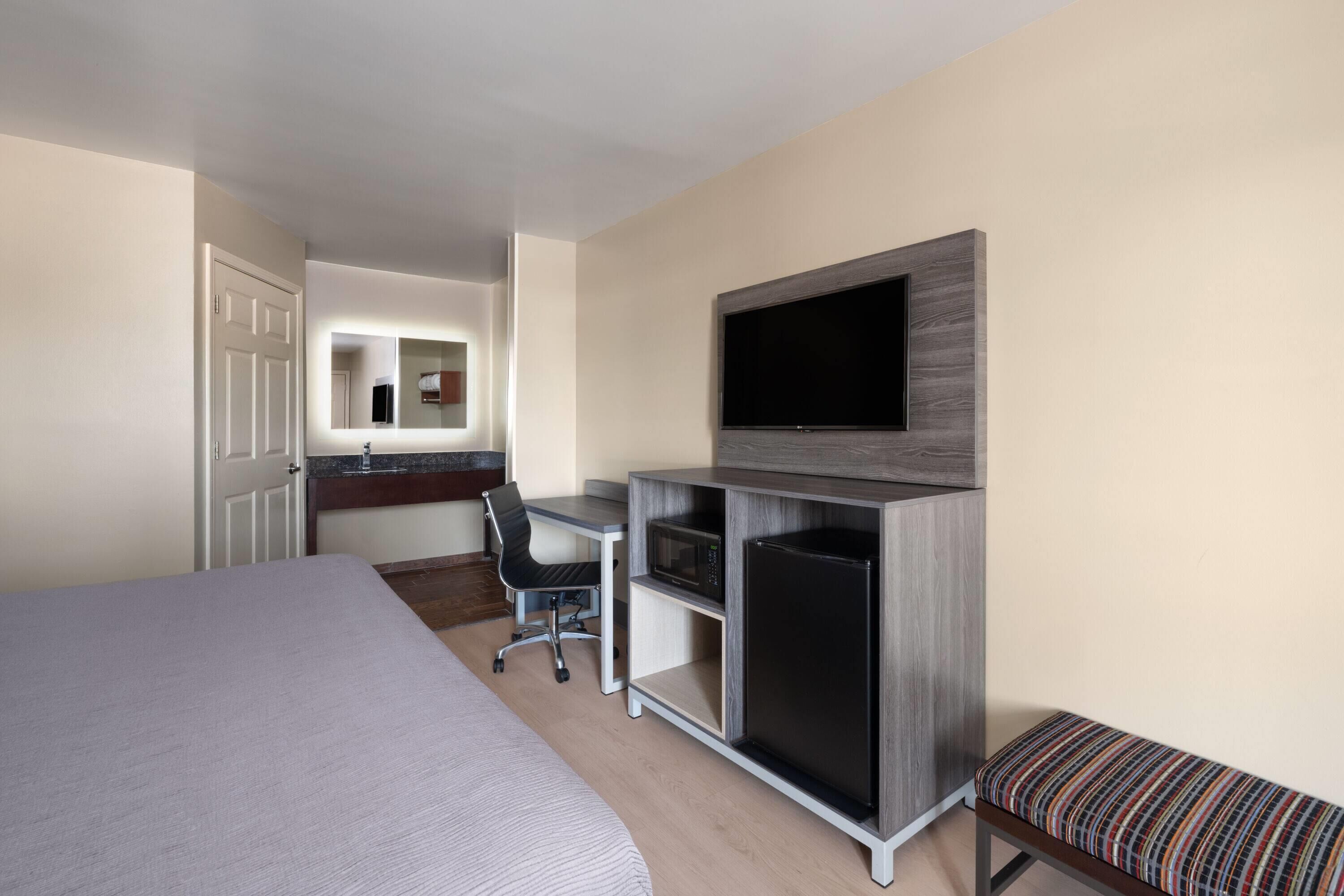 Habitación de hotel con una cama grande, un escritorio con silla, un televisor en la pared y un refrigerador pequeño.
