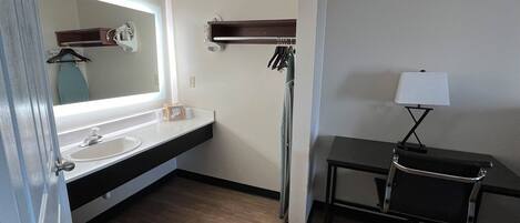 A hotel bathroom with a sink, mirror, towel rack, and a desk with a lamp.