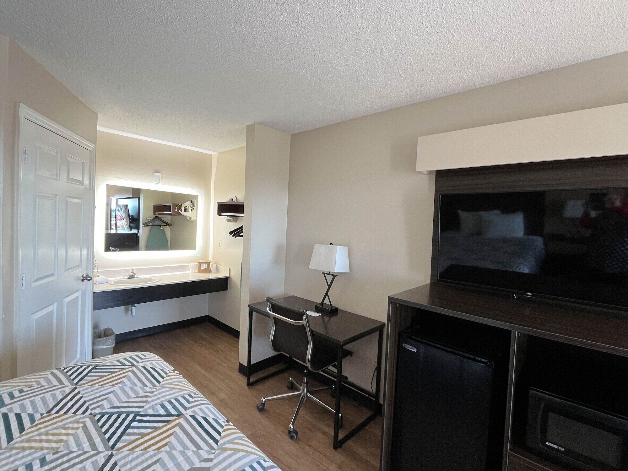 Habitación de hotel con una cama, un escritorio con silla, televisión, espejo, lavamanos y una pequeña mesa.