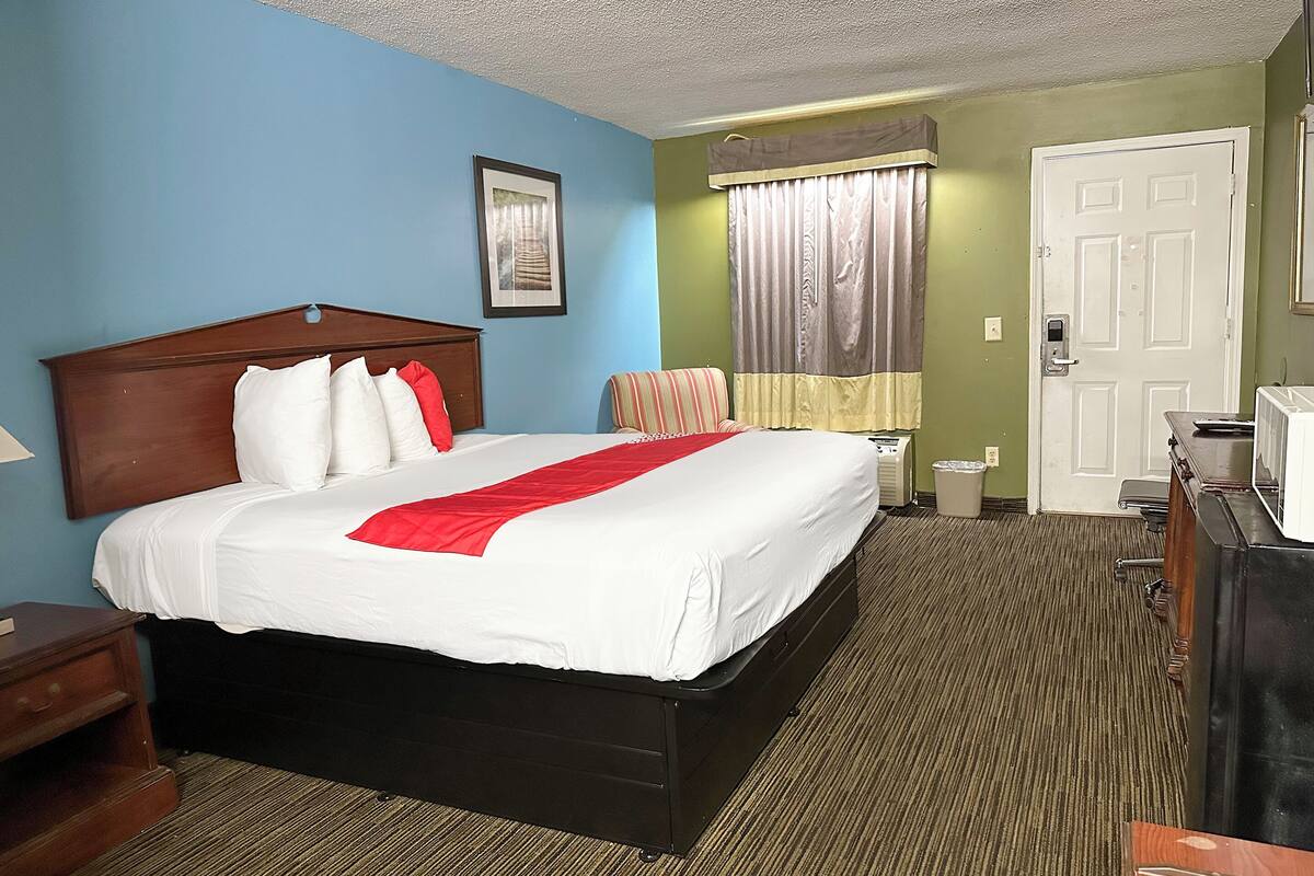 A hotel room with a large bed, a wooden headboard, a nightstand, a desk with a chair, a window with curtains, and a door.
