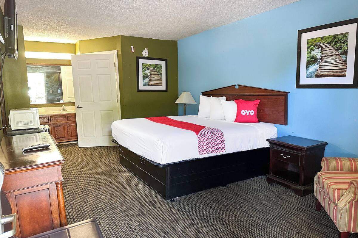 A hotel room with a bed, bedside table, lamp, and a red pillow.