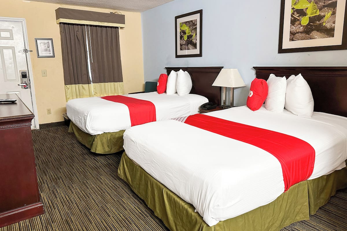 A hotel room with two beds, each with white bedding and red accents, a wooden headboard, a desk with a chair, and a window with curtains.