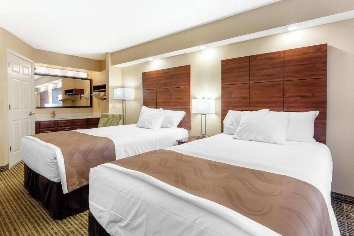 Ein Hotelzimmer mit zwei Betten, einem großen Spiegel, einem Schminktisch mit Waschbecken und einem Kopfteil mit integrierter Beleuchtung.