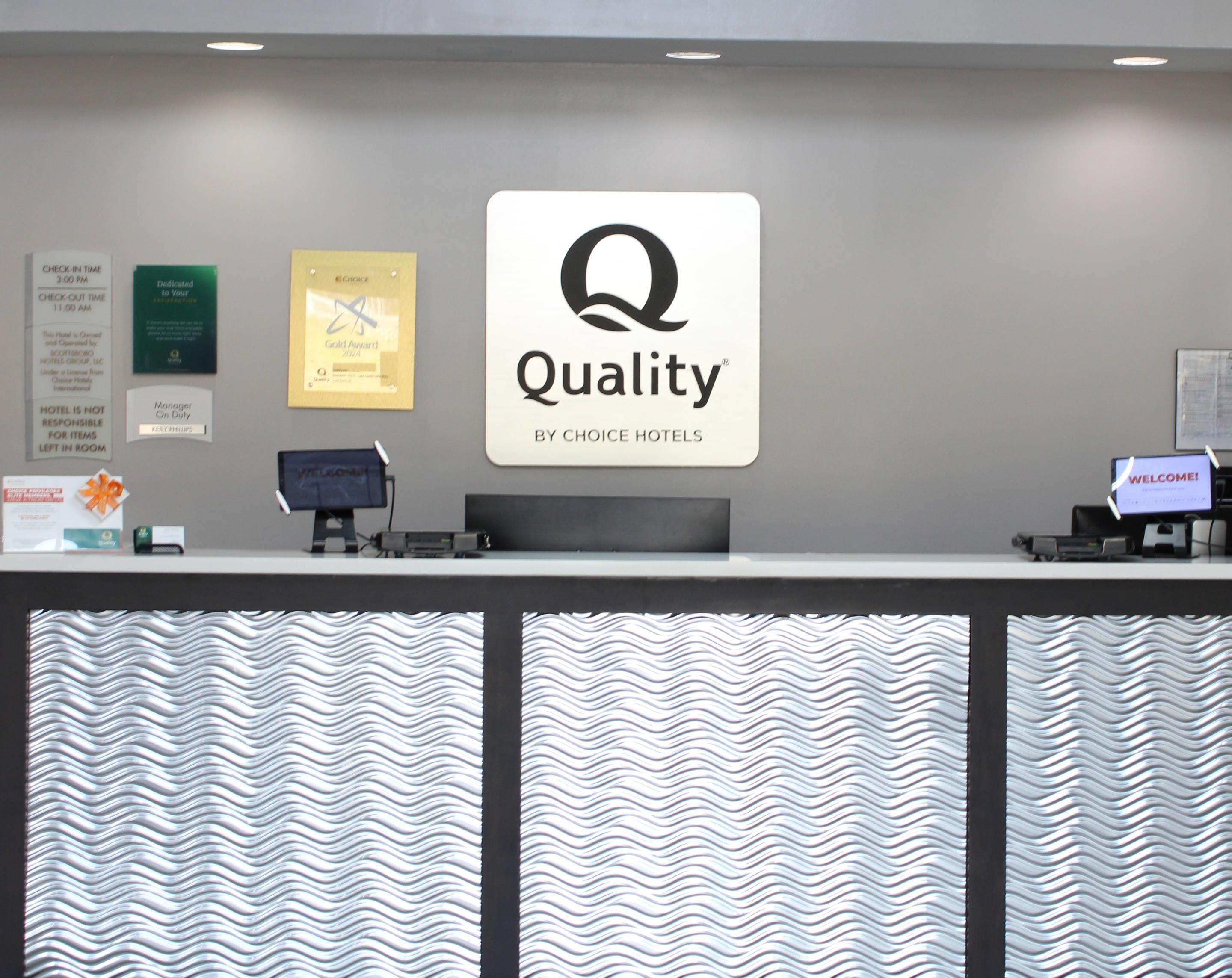 A hotel reception desk with a large 'Quality by Choice Hotels' sign, a 'Manager On Duty' sign, and a 'Welcome!' sign.