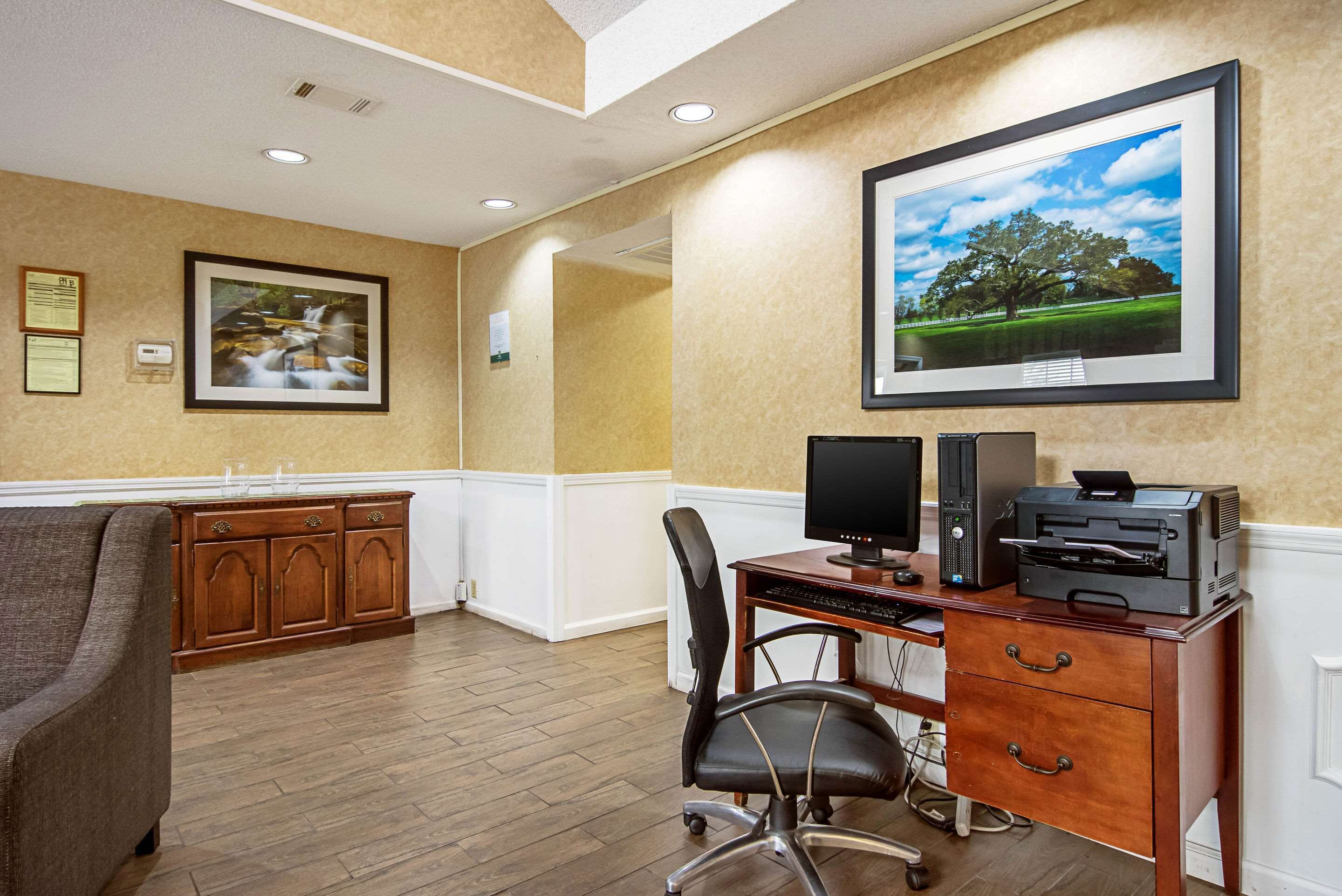 A home office with a desk, computer, printer, and a chair. There are framed pictures on the wall and a cabinet with drawers.
