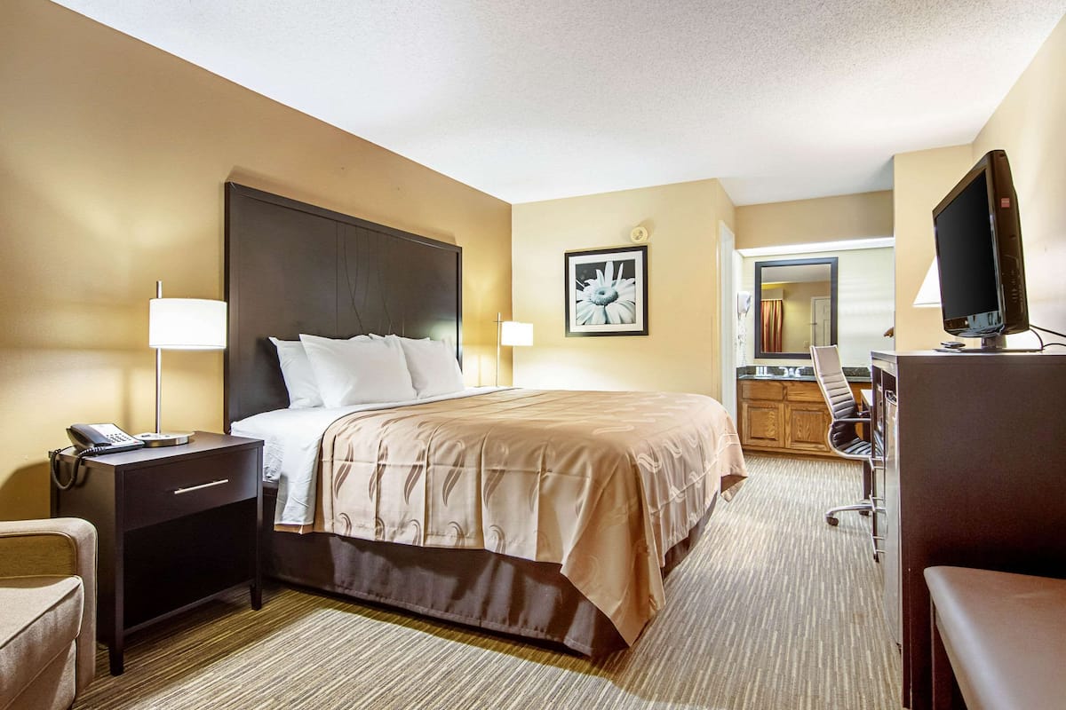 A hotel room with a bed, bedside table, lamp, desk with a computer, and a TV.