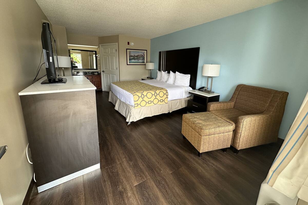 Una habitación de hotel con una cama, un escritorio con computadora, una silla y una mesita con lámpara.