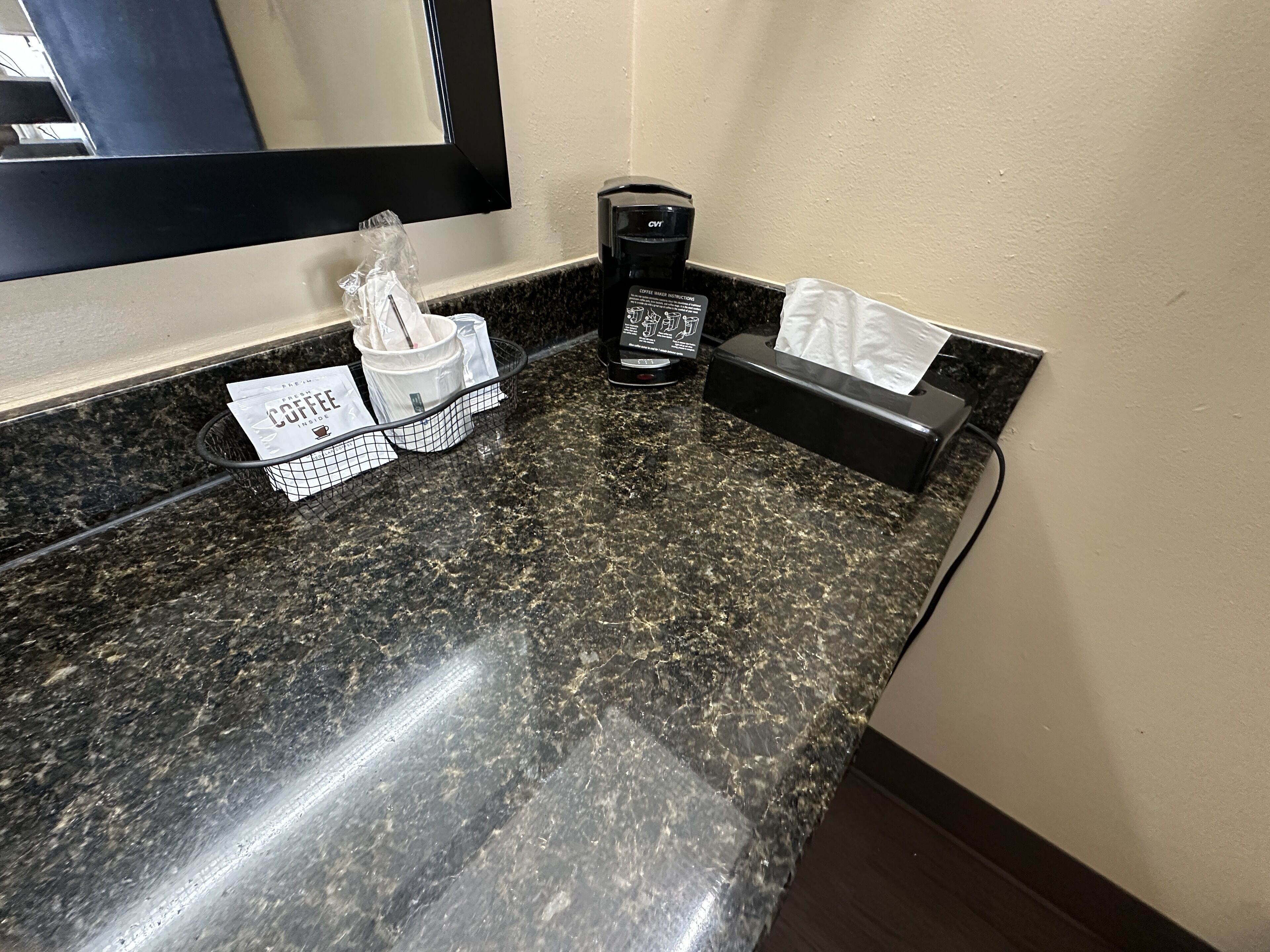 A hotel bathroom counter with a coffee maker, a tissue box, and a paper towel dispenser.