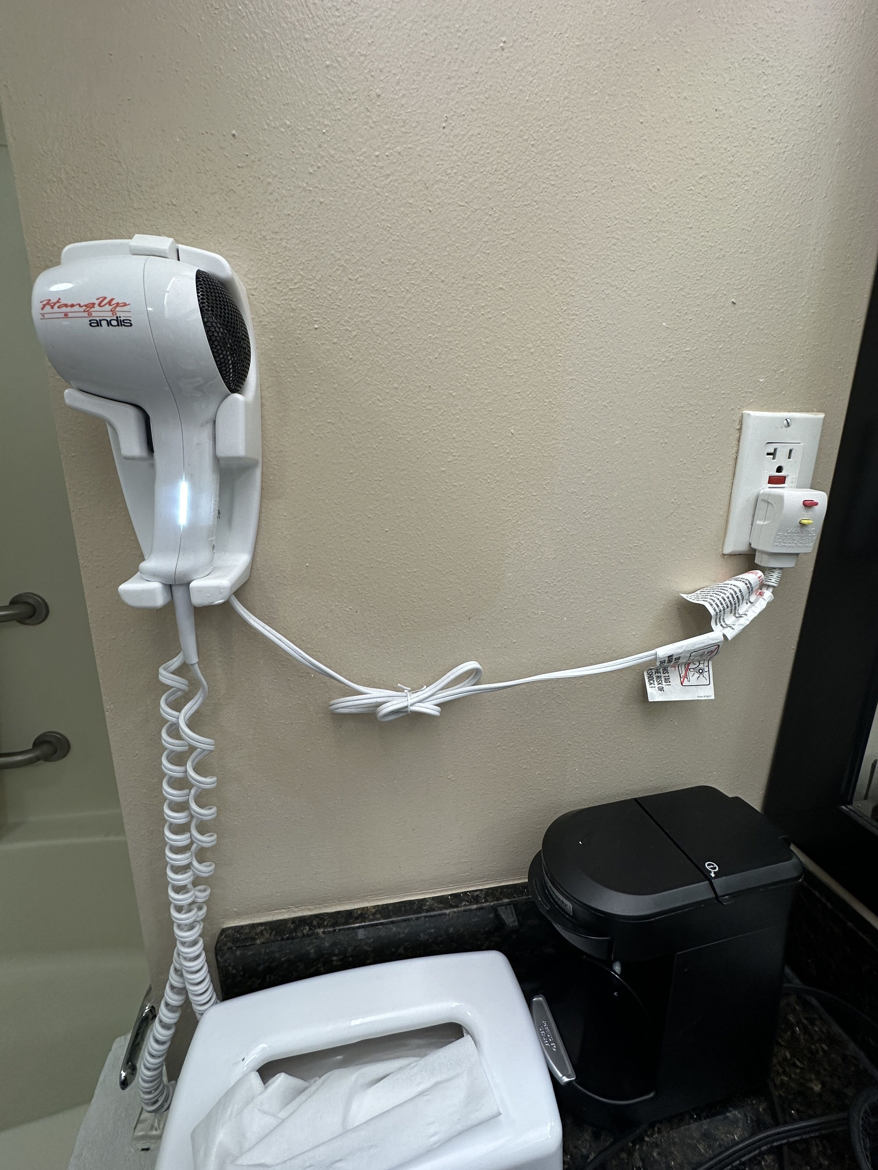 Un sèche-cheveux fixé au mur, branché à une prise de courant, avec des toilettes et une poubelle à proximité.