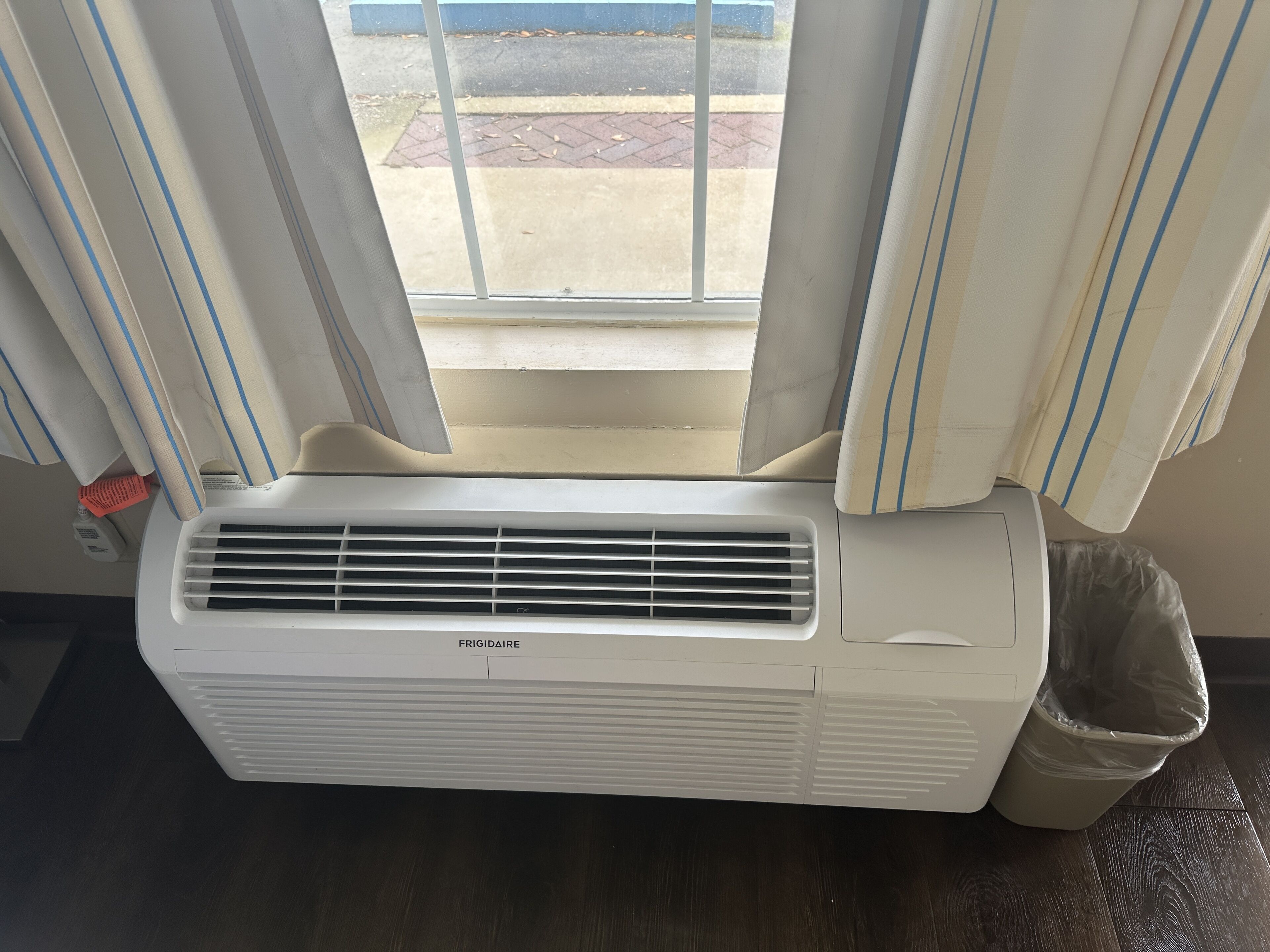 A white air conditioner unit labeled "FRIGIDAIRE" in a room with a window, curtains, and a trash bin.