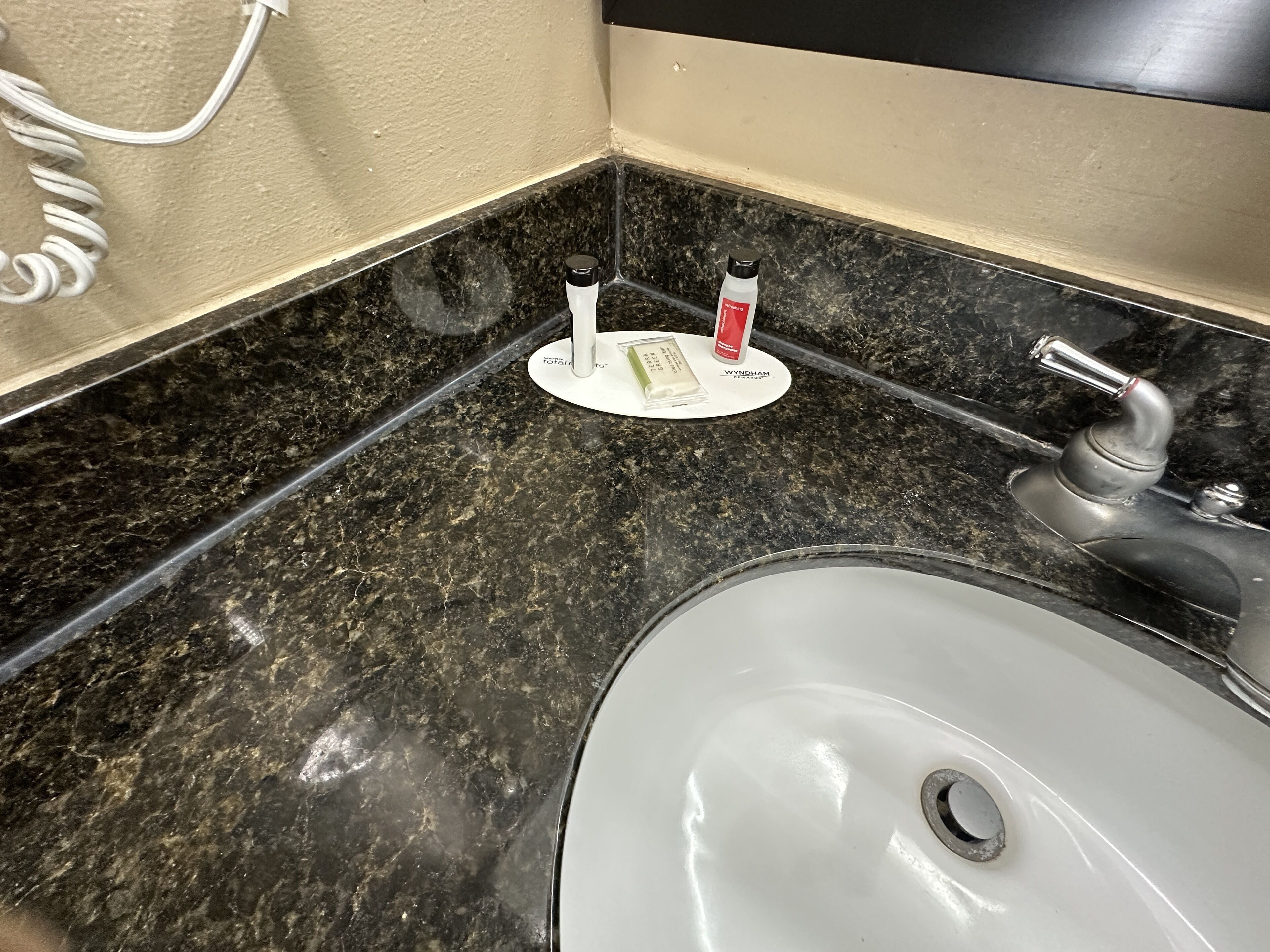 Une salle de bain avec un lavabo blanc, un plan de toilette en marbre noir et un distributeur de serviettes blanches sur lequel repose une bouteille rouge.