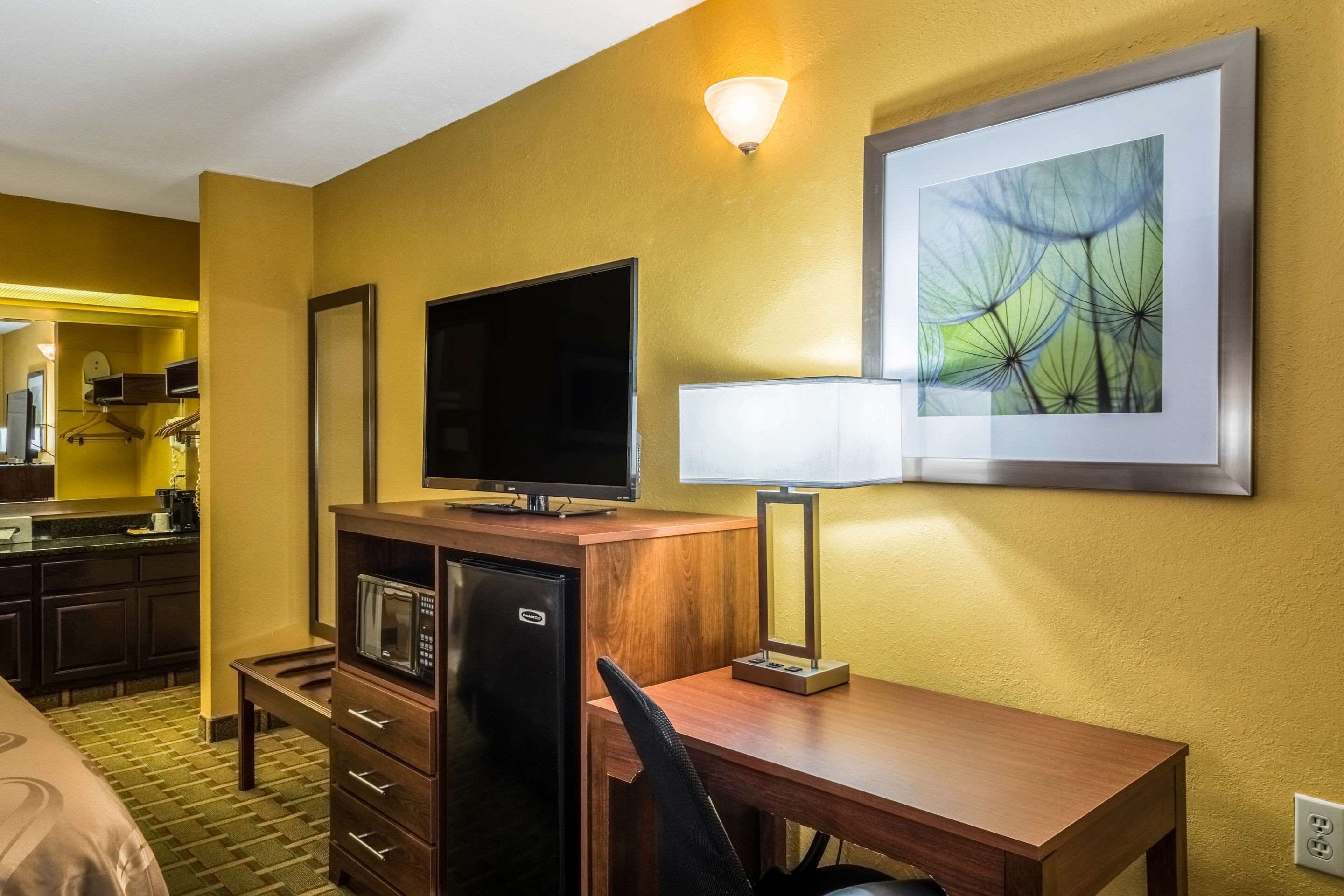 Una habitación de hotel con un escritorio de madera, televisión, microondas, lámpara, una obra de arte enmarcada y una kitchenette con cafetera.