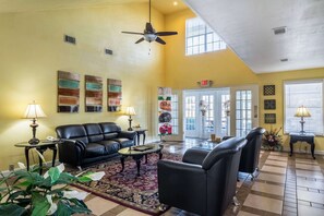 A spacious living room with a black leather sofa, a coffee table, and a ceiling fan. There are two lamps on side tables, a large rug, and a door leading outside.