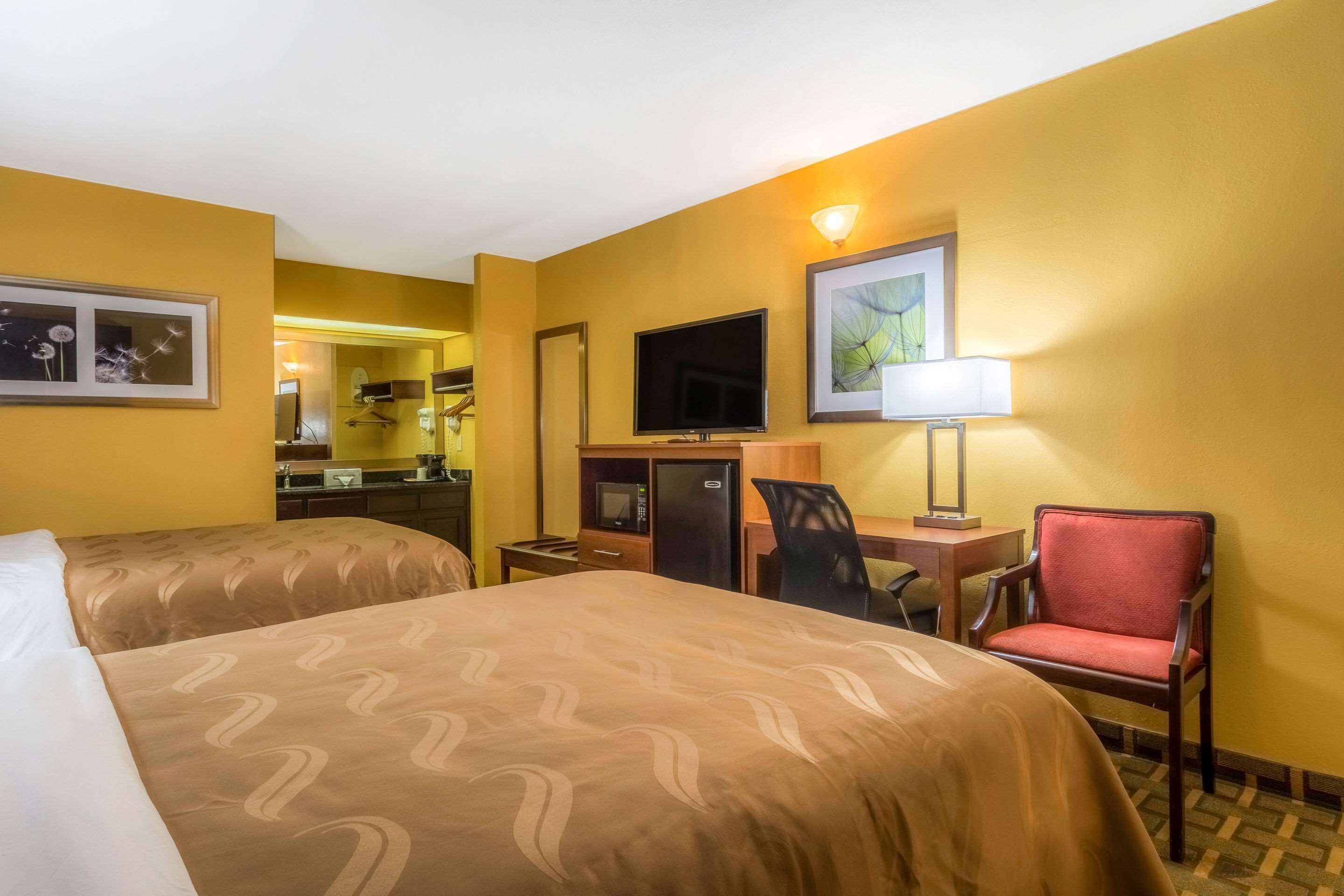 Ein Hotelzimmer mit zwei Betten, einem Schreibtisch mit Stuhl, einem Fernseher, einer Mikrowelle und einer kleinen Küchenzeile.