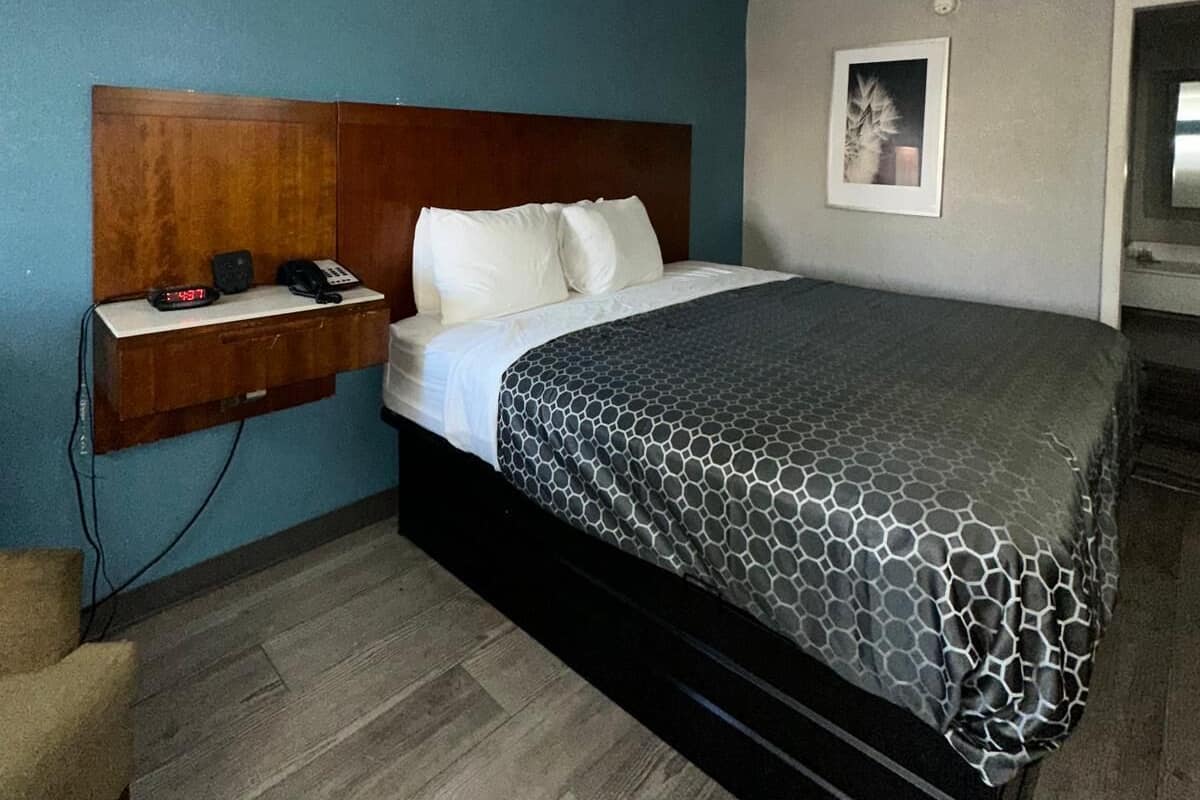 A hotel room with a bed, a wooden headboard, a bedside table with a phone and a clock, a picture on the wall, and a wooden floor.