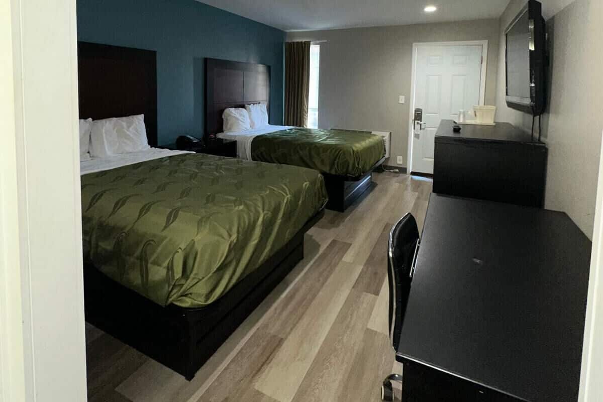 A hotel room with two beds, a desk, a chair, a TV, and a door.