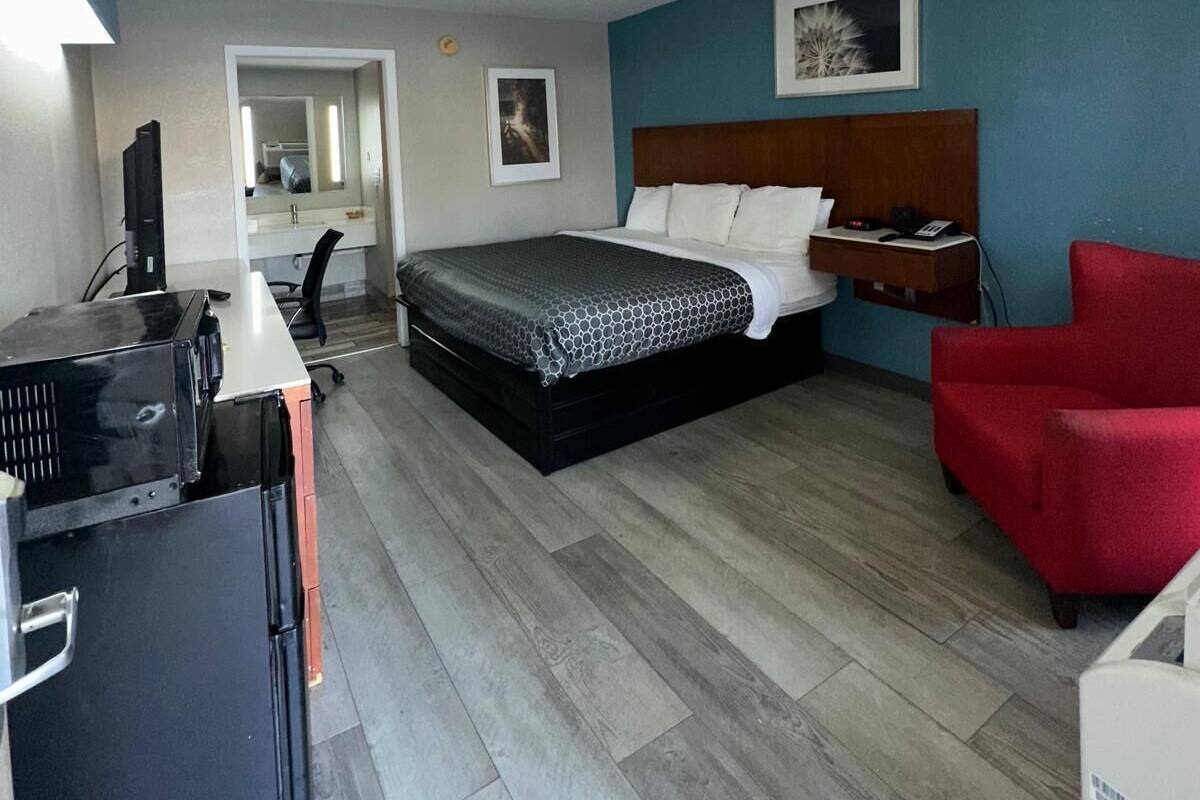 A hotel room with a bed, a desk with a computer, a red chair, and a small table with a phone and a lamp.