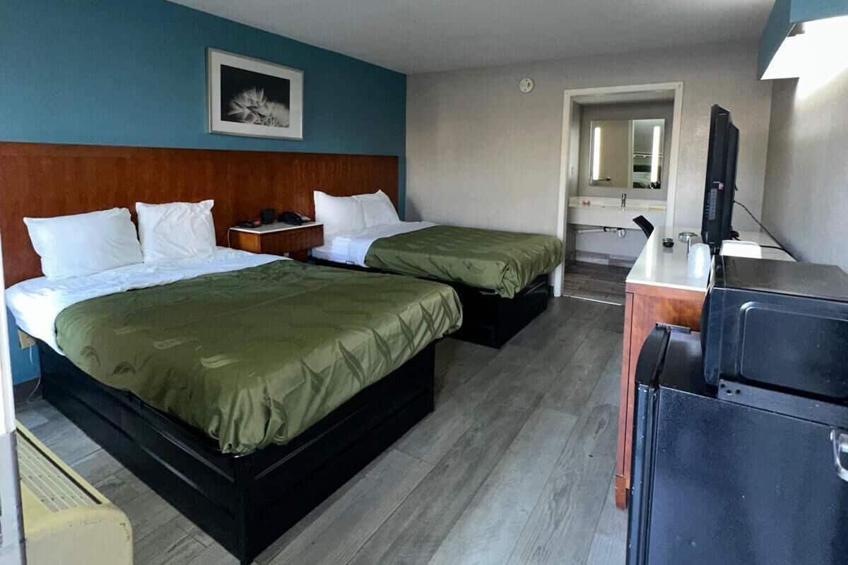 A hotel room with two beds, a desk, a refrigerator, and a bathroom visible through an open door.
