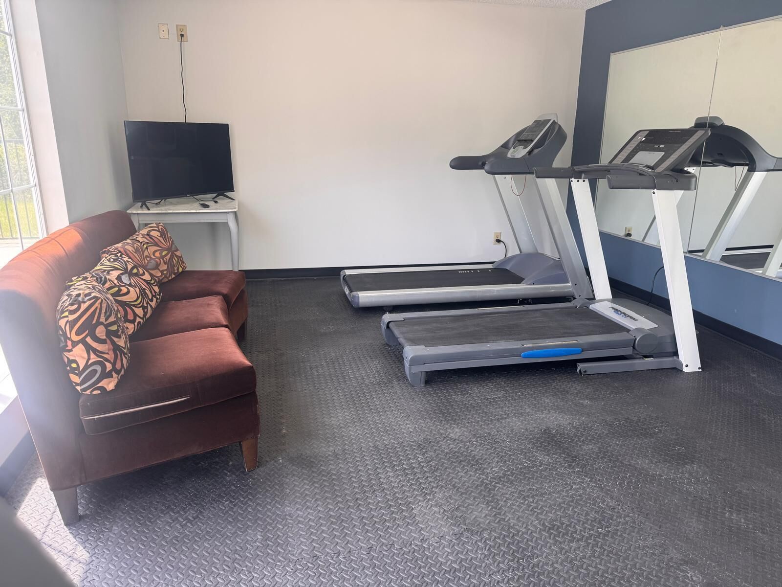 Une pièce comprenant un tapis de course, un canapé avec des coussins à motifs, un téléviseur posé sur un meuble et une fenêtre.
