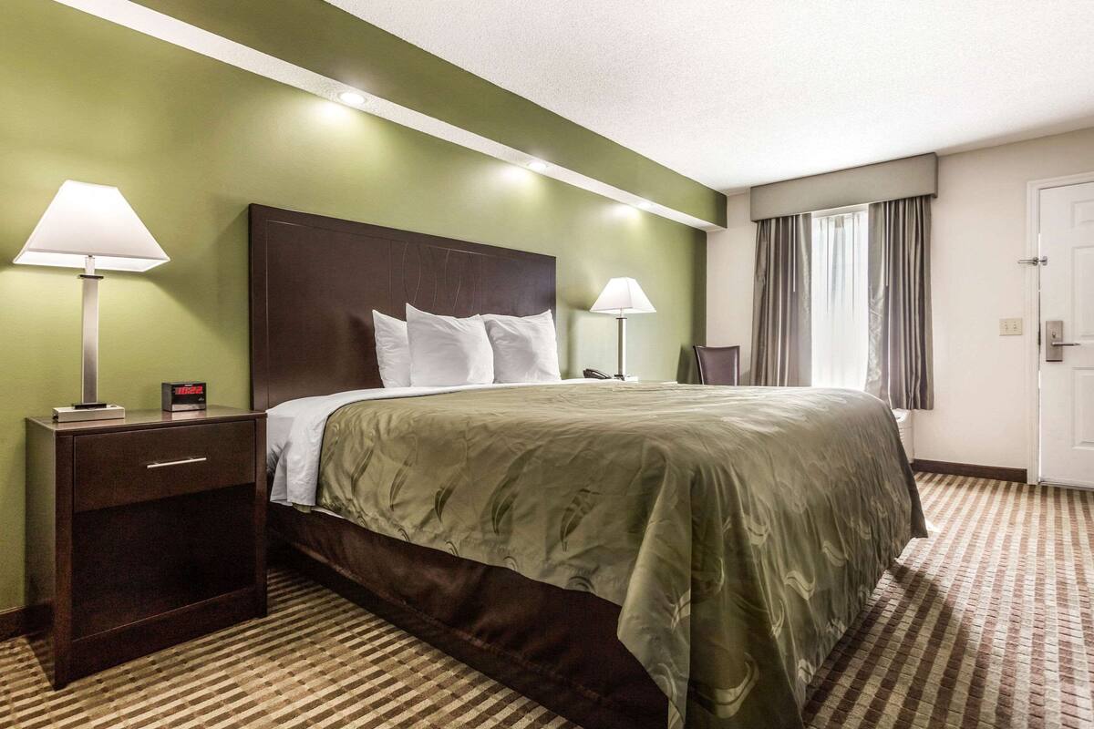 A hotel room with a large bed, two bedside tables with lamps, a nightstand, a window with curtains, and a door.