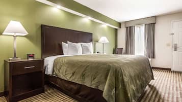 Una habitación de hotel con una cama grande, dos mesitas de noche con lámparas, una mesita auxiliar, una ventana con cortinas y una puerta.