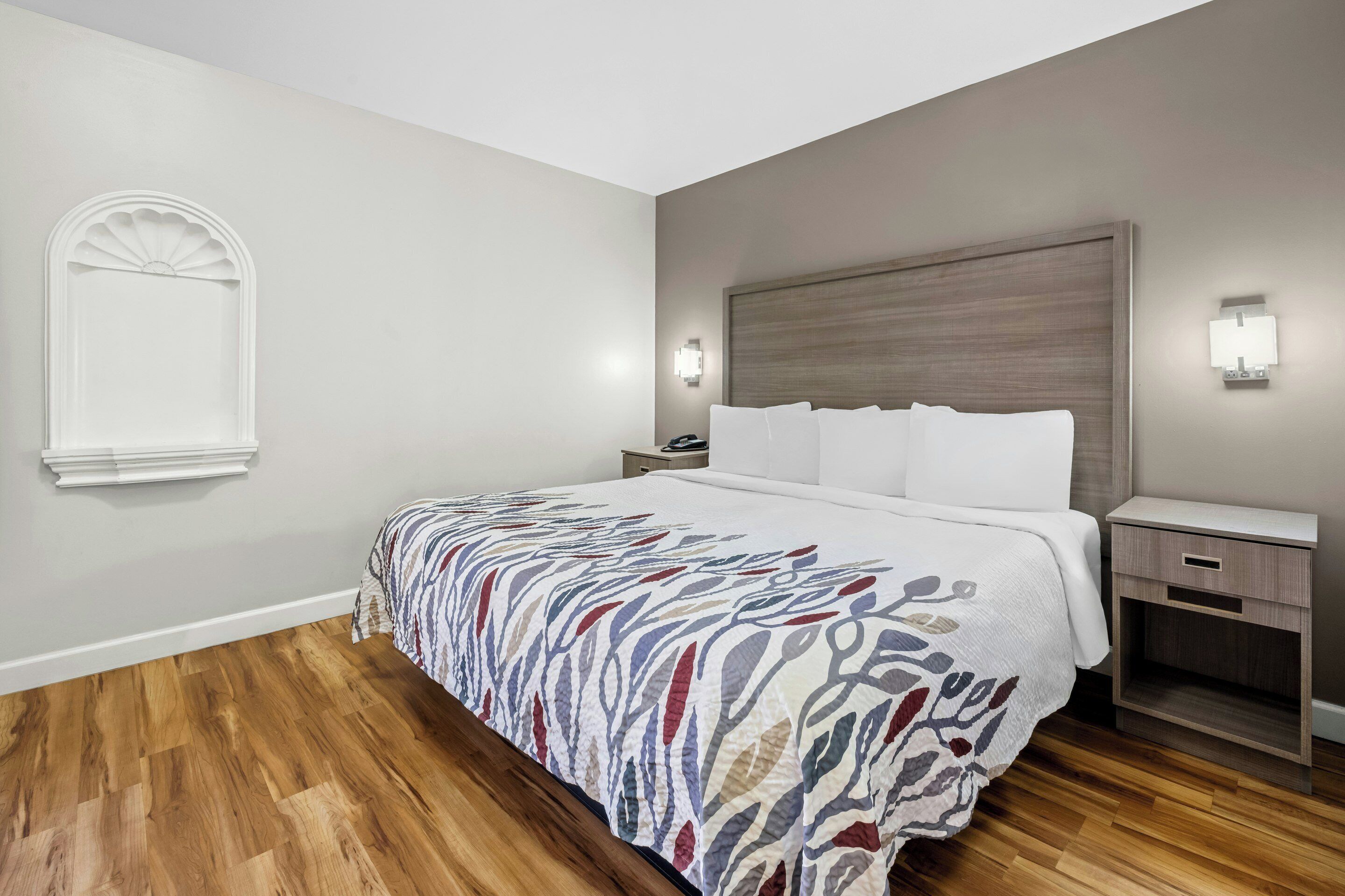 Habitación de hotel con cama, mesitas de noche, una lámpara de pared y una ventana con detalles decorativos.