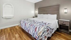 A hotel room with a bed, bedside tables, a nightstand, a wall-mounted light, and a window with a decorative trim. - Red Roof Inn & Suites Wilson (Wilson)