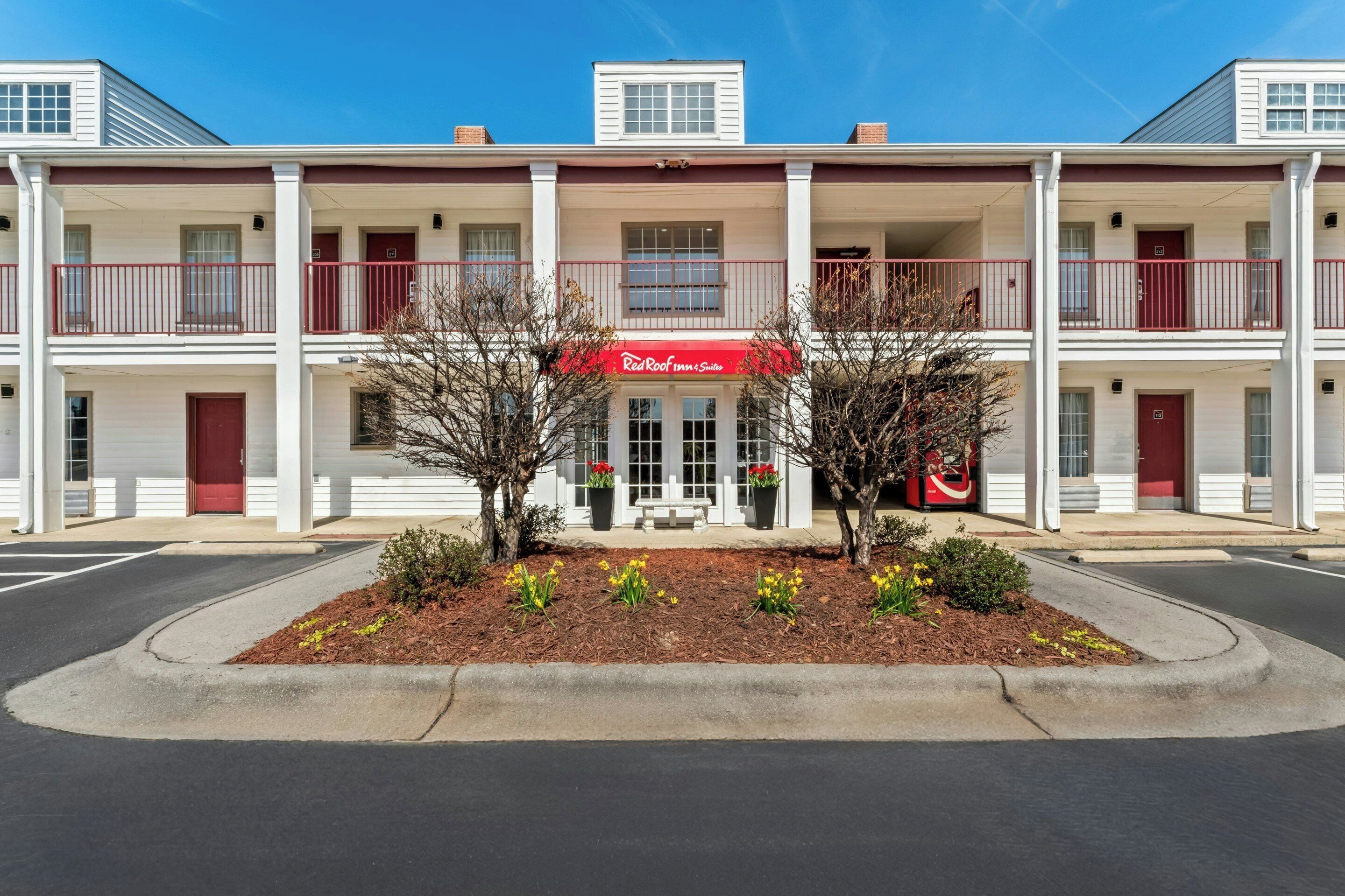 Ein zweistöckiges Motel mit roter Markise, einer überdachten Veranda und einem begrünten Vorgarten mit kleinen Bäumen und Blumen.