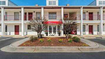 Ein zweistöckiges Motel mit roter Markise, einer überdachten Veranda und einem begrünten Vorgarten mit kleinen Bäumen und Blumen.