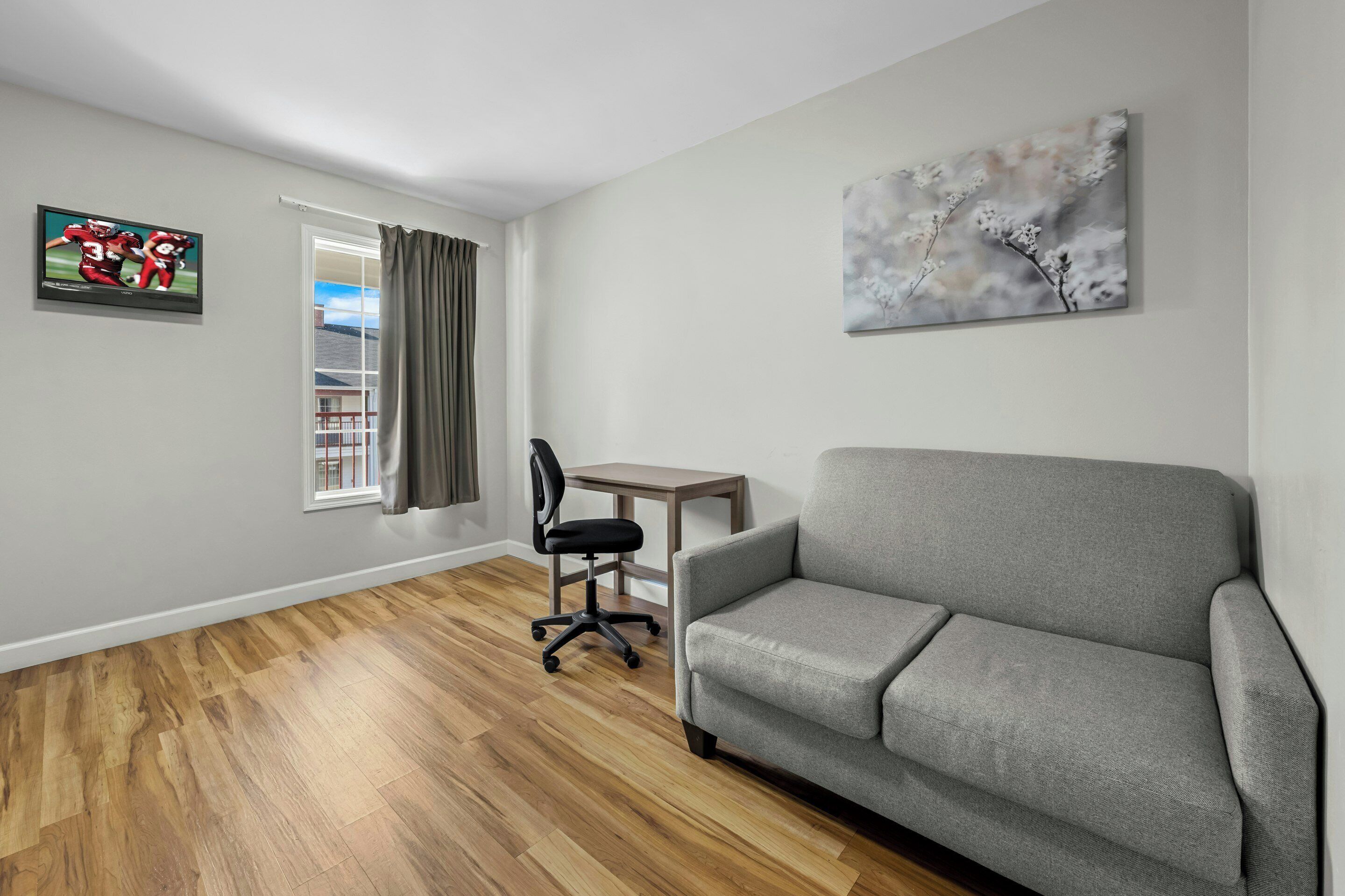 Una sala de estar con un sofá gris, suelo de madera, un televisor en la pared, una mesita y una silla.