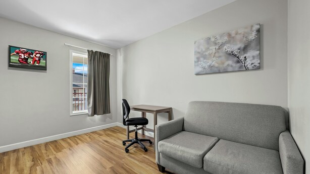 A living room with a grey sofa, wooden flooring, a TV on the wall, a small table, and a chair. - Red Roof Inn & Suites Wilson (Wilson)