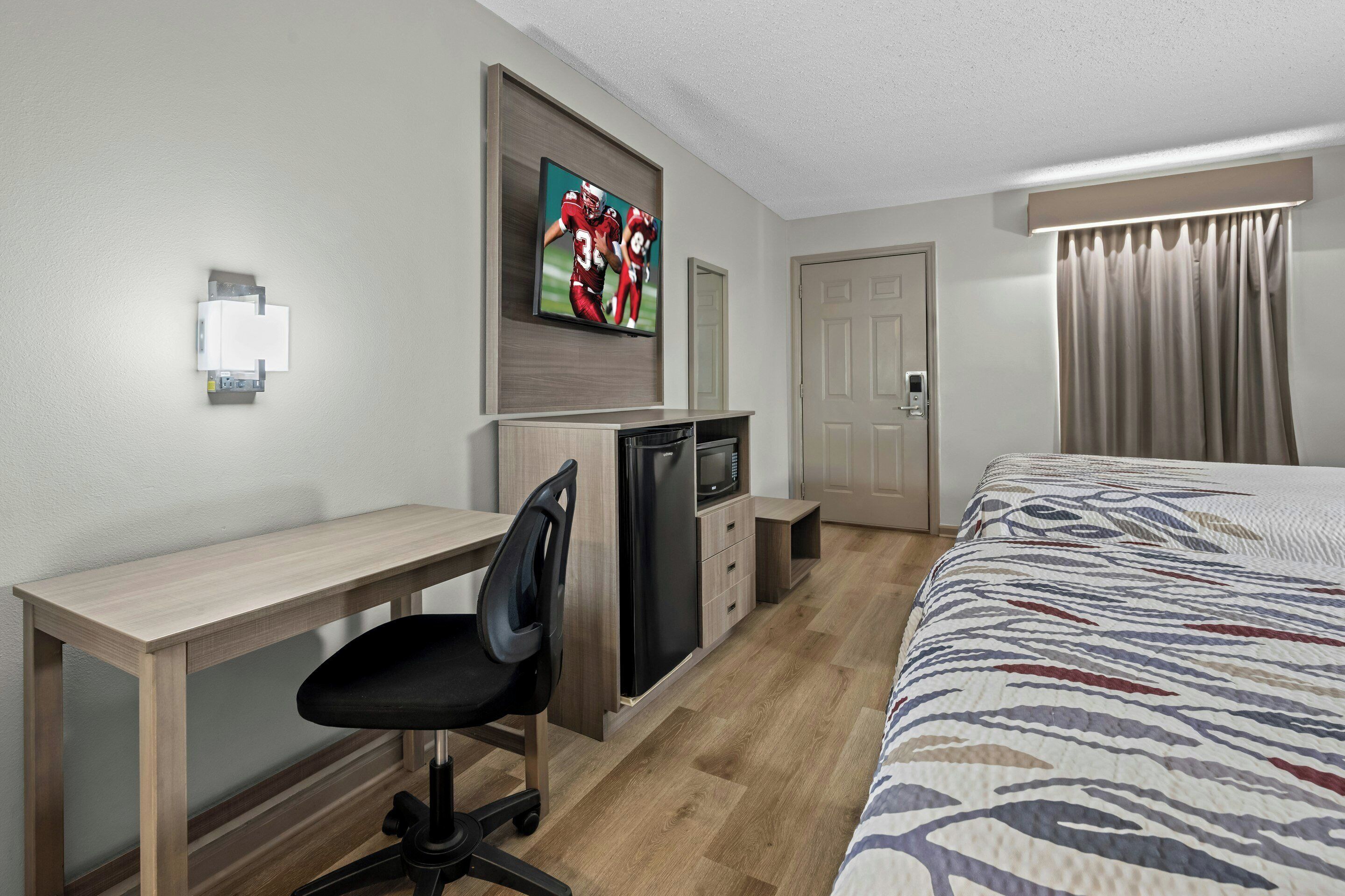 A hotel room with a wooden desk, a black office chair, a bed with a patterned bedspread, a television mounted on the wall, a microwave, and a refrigerator.