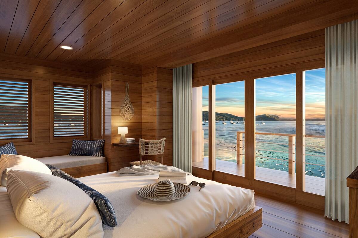 Ein Holzhüttenraum mit einem Bett, einem Sessel, einer Lampe und Blick auf das Meer.