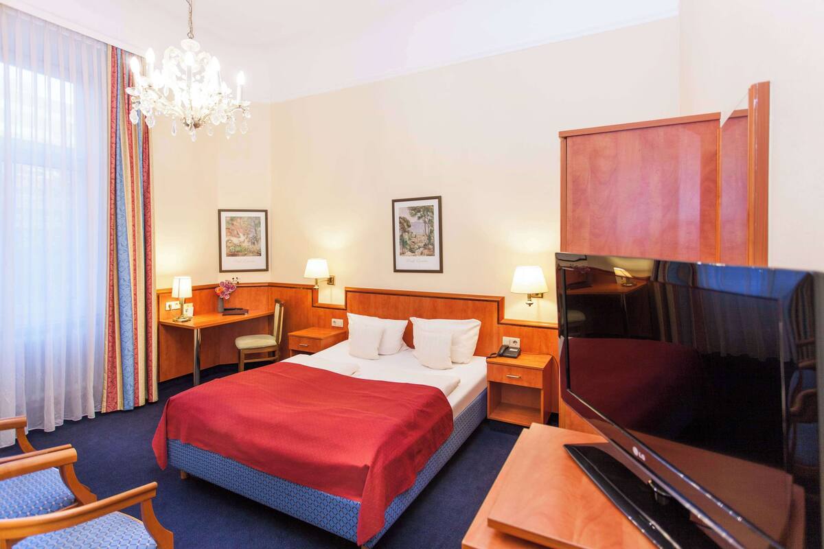 Une chambre d’hôtel avec un lit, un bureau, une chaise, une télévision et un lustre.