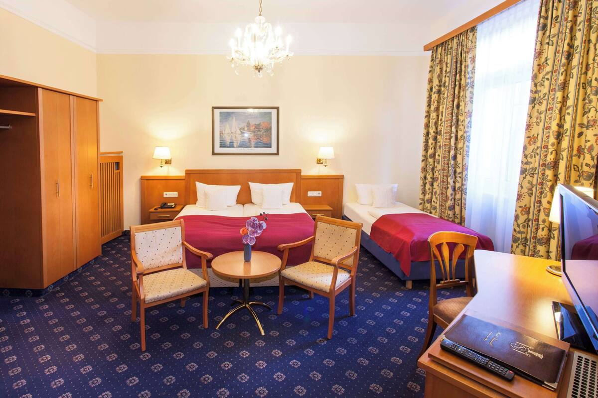Une chambre d’hôtel comprenant un lit, deux tables de chevet avec des lampes, un bureau avec une chaise, une télévision et un lustre.