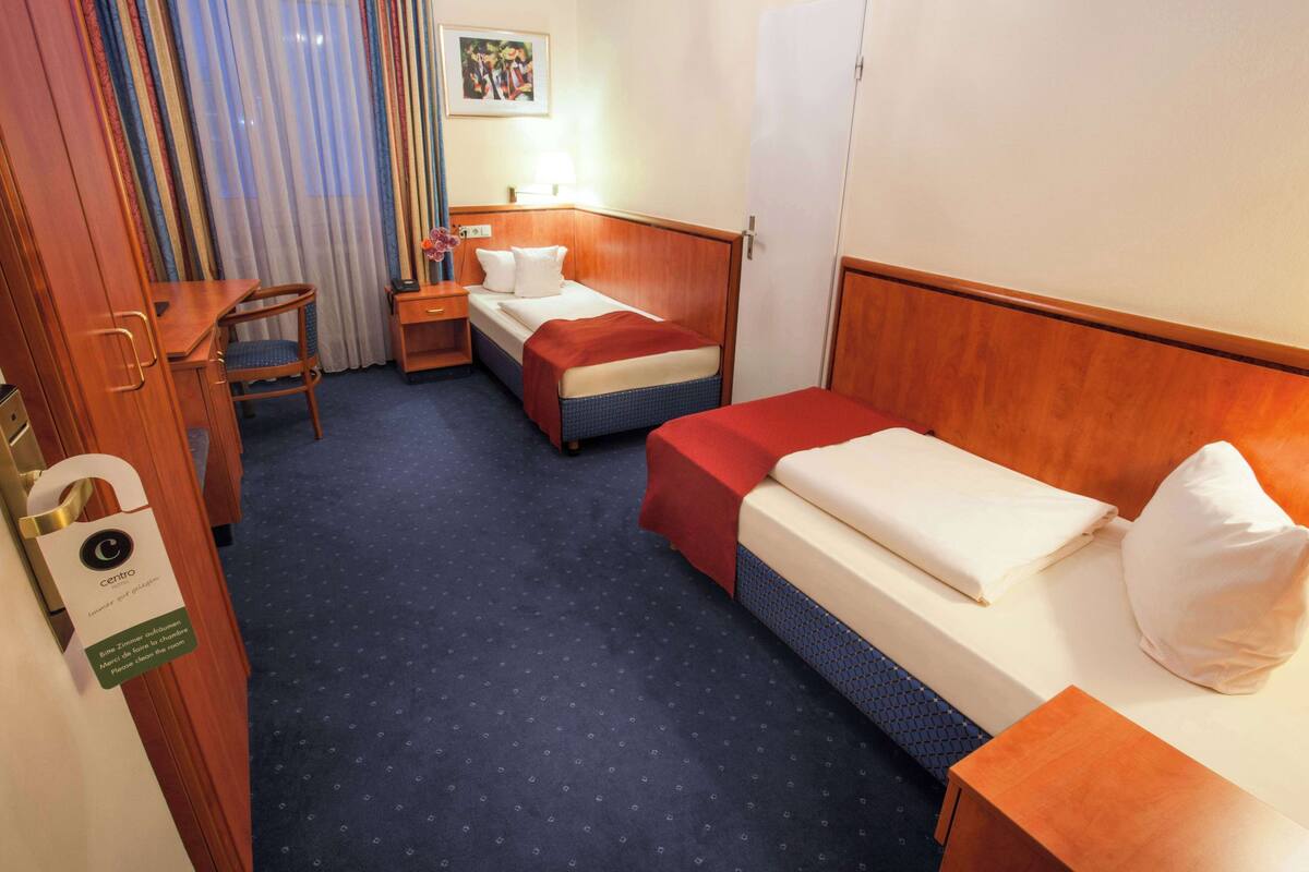 Une chambre d’hôtel avec un lit, un bureau, une chaise, une table de chevet et une fenêtre avec des rideaux.