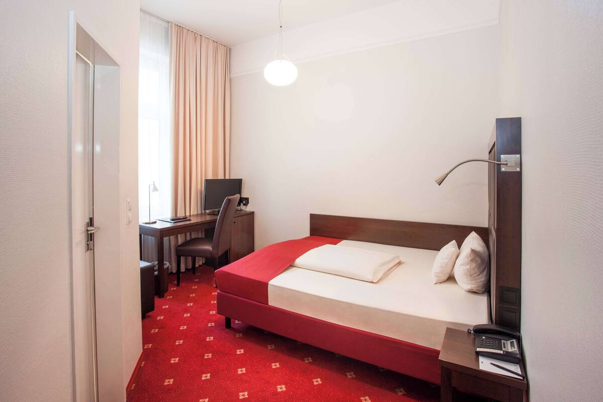 Une chambre d’hôtel comprenant un lit, un bureau avec une chaise, une lampe de bureau, un téléphone et une fenêtre avec des rideaux.