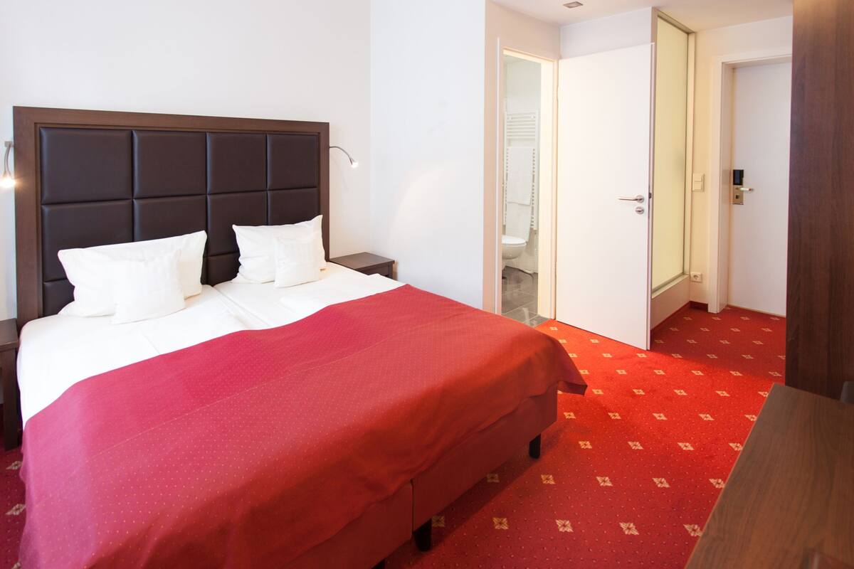 Une chambre d’hôtel avec un grand lit, une table de chevet, un sol recouvert d’une moquette rouge et une porte donnant sur une salle de bain.