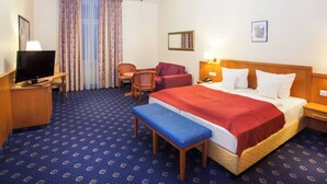 Una habitación de hotel con una cama, un escritorio con televisor, una silla, un banco y una araña.