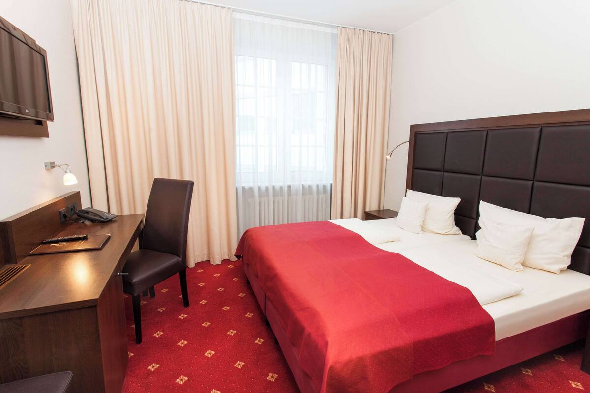 Une chambre d’hôtel avec un lit, un bureau, une chaise, une télévision et une fenêtre avec des rideaux.