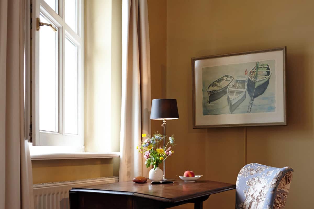 A hotel room with a bed, a desk with a lamp, a chair, a window with curtains, and a painting on the wall.