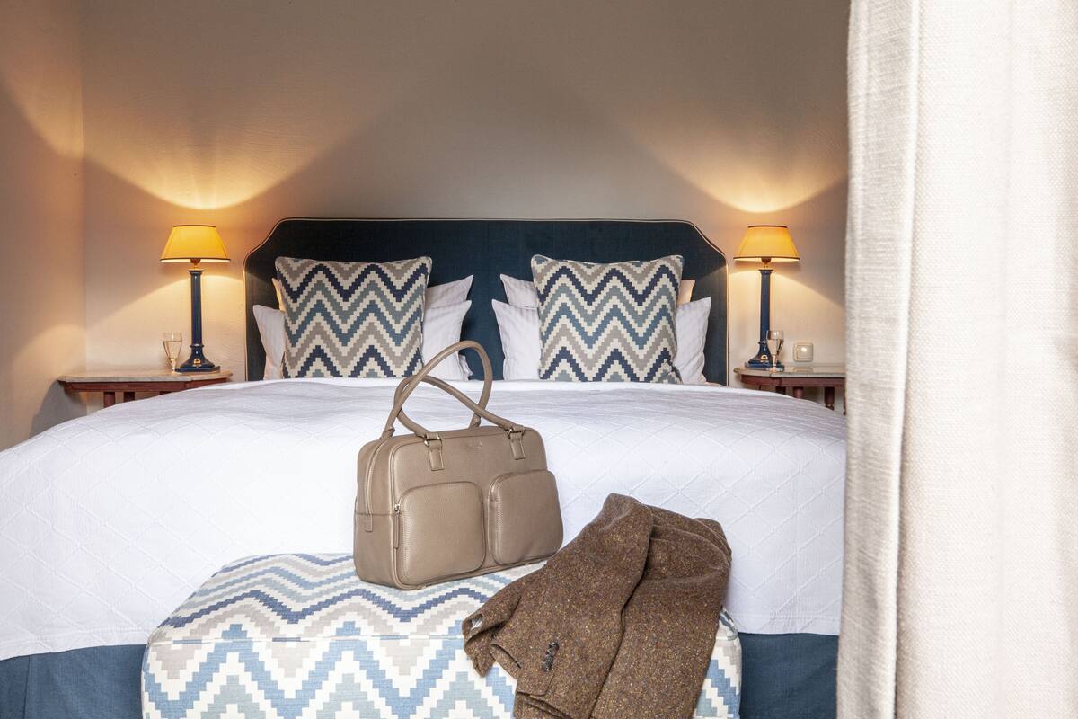 A neatly made bed with a patterned bedspread, two pillows, a handbag, and a jacket on the bed.