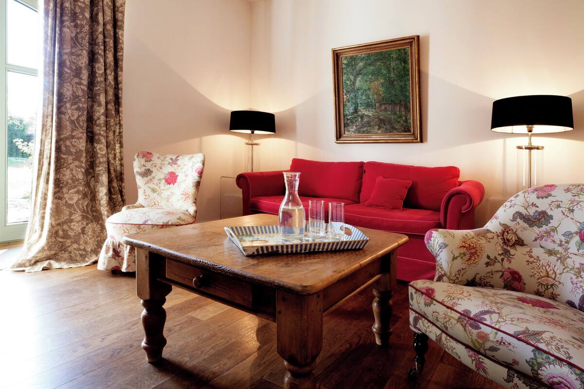 A living room with a wooden coffee table, floral armchairs, a red sofa, and two floor lamps.
