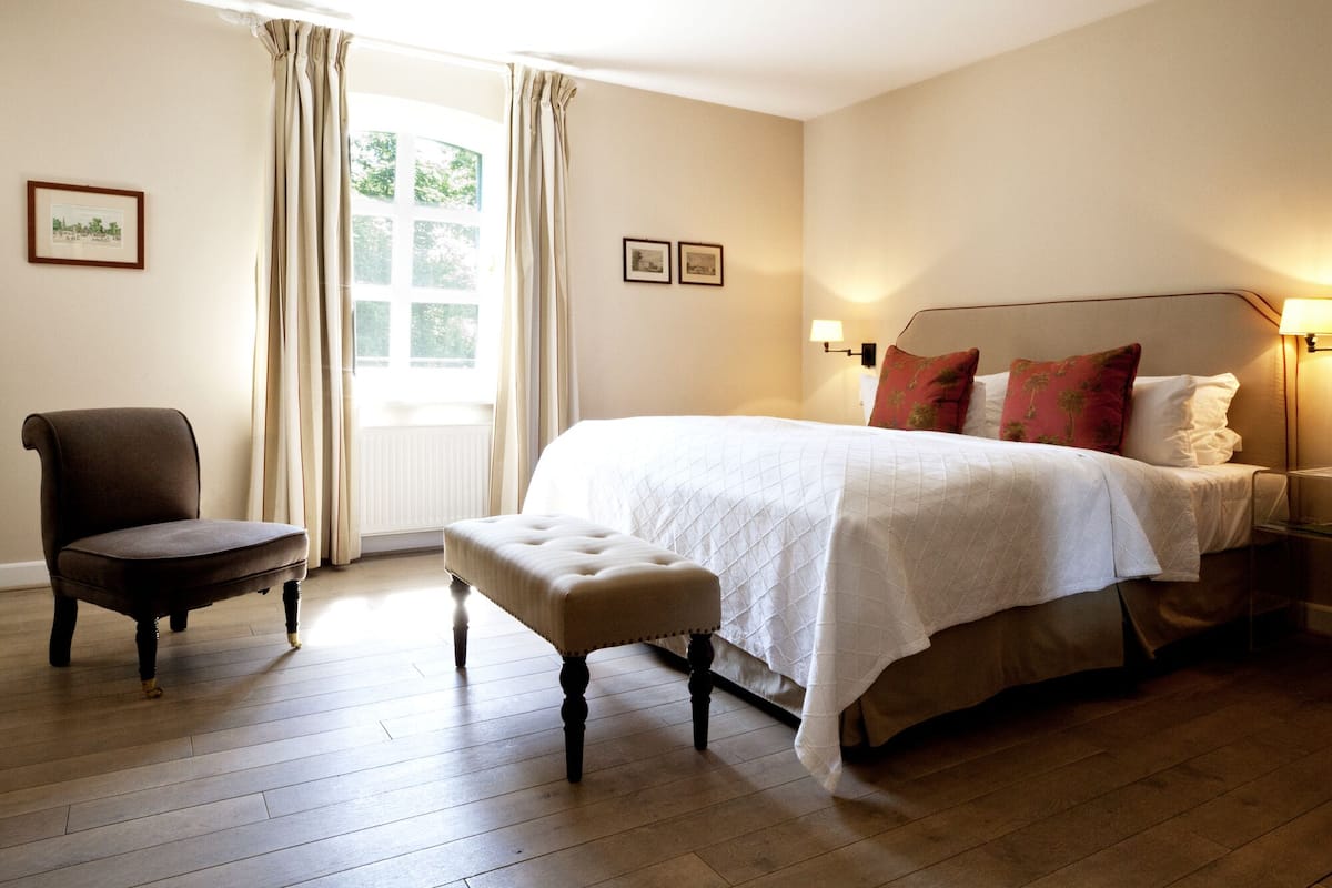 A hotel room with a large bed, a chair, an ottoman, and a window with curtains.