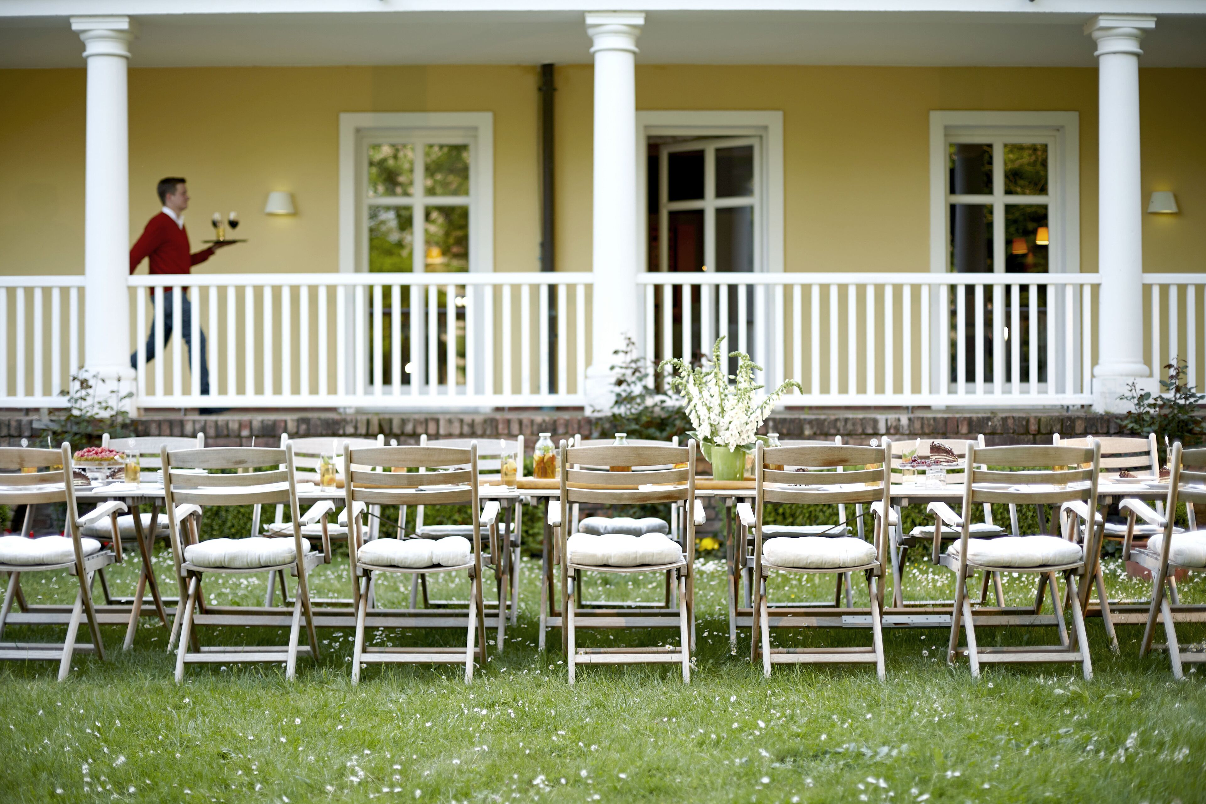 A covered outdoor dining area with multiple tables and chairs, a waiter in the background, and a yellow building with white columns.