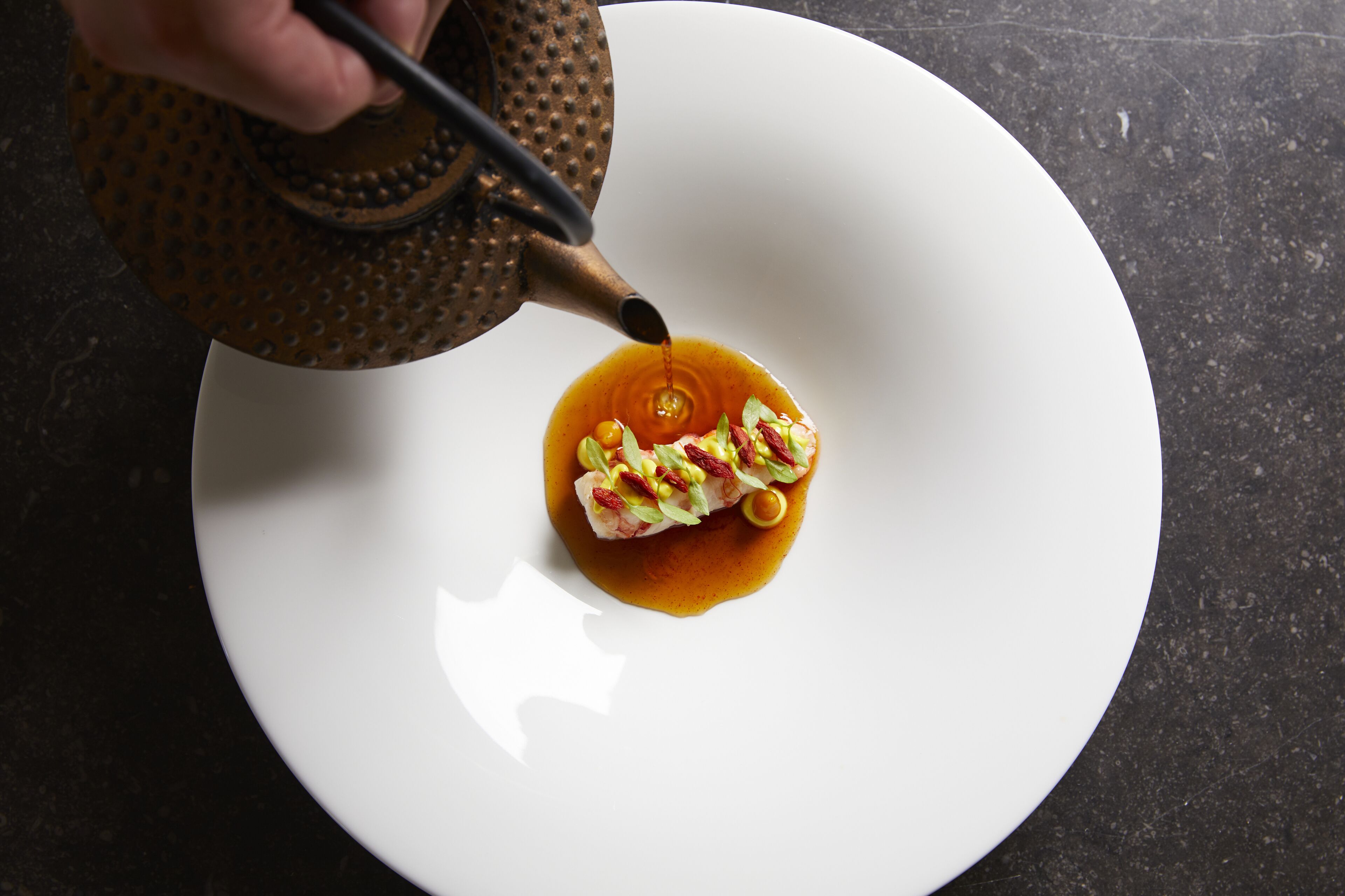 A white plate with a food item and a teapot pouring liquid onto it.