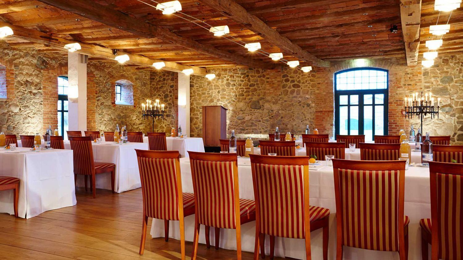 A restaurant with stone walls, wooden beams, and striped chairs. Tables are set with white tablecloths and candles.