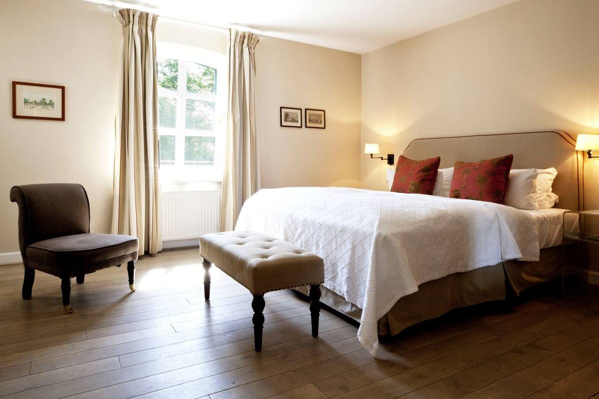 A hotel room with a bed, a chair, a bench, a window with curtains, and wall-mounted lamps.