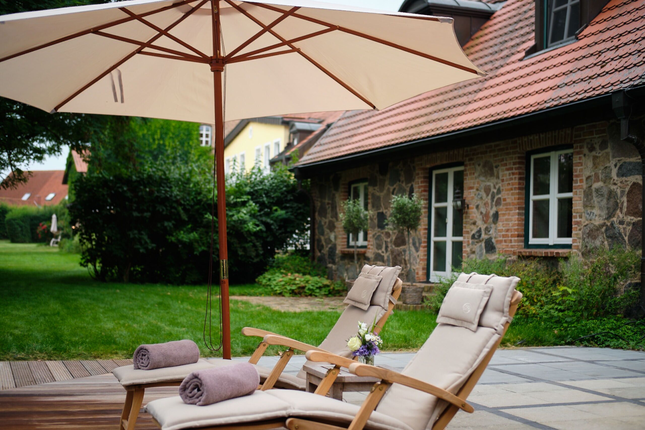 A patio with two lounge chairs, a beige umbrella, and a stone building in the background.