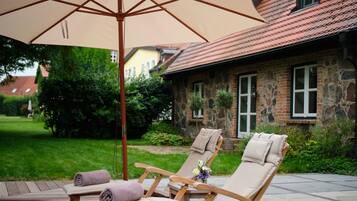 A patio with two lounge chairs, a beige umbrella, and a stone building in the background.